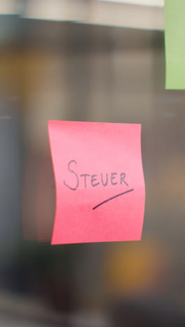 Vertical-Video-Close-Up-Of-Woman-Putting-Sticky-Note-With-German-Word-Steuer-Or-Tax-Written-On-It-Onto-Transparent-Screen-In-Office-